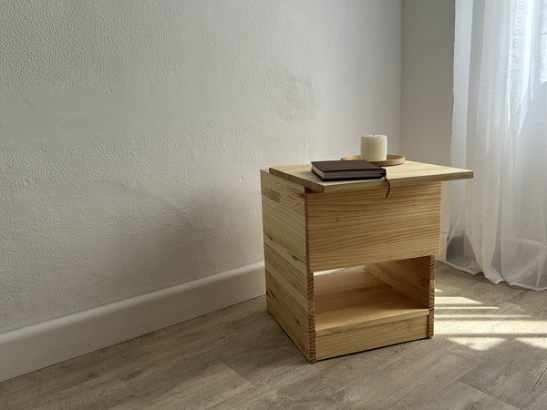 Mesita de noche Nocternum de pino natural junto a una ventana, con libreta, vela y bandeja encima